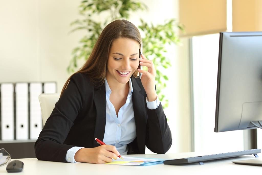 Smiling person on a cell phone call sitting in front of a computer and writing.